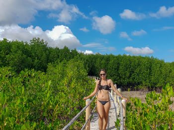 Portrait of woman in bikini standing on boardwalk against sky