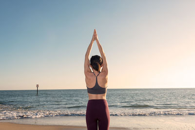 Back view of unrecognizable female in activewear standing in mountain pose with arms up while practicing yoga on sandy beach near waving sea