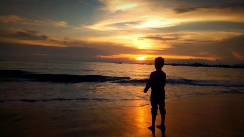 Silhouette boy standing at beach against sky during sunset