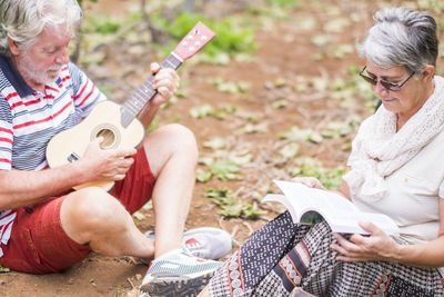 Senior man playing ukulele with woman reading book on field