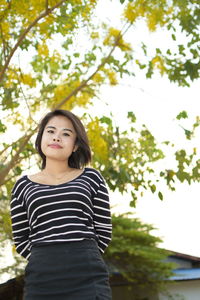 Low angle portrait of beautiful young woman standing against trees in park