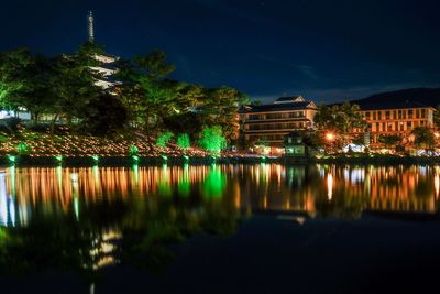 Illuminated buildings by lake against sky at night