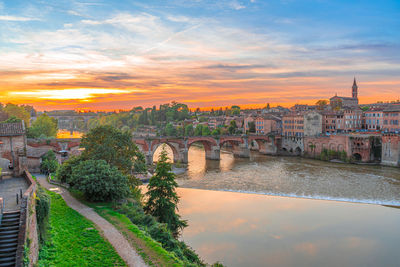 Bridge over river by buildings against sky during sunset