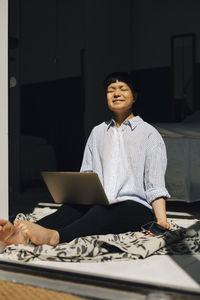 Smiling woman with laptop sitting in sunlight at home