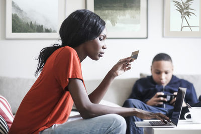 Mother holding credit card while using laptop with son in background at home