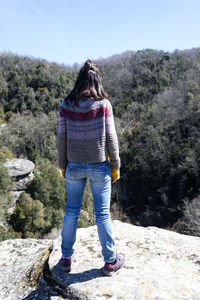 Back view of traveler woman in sweater at mountains against forest.