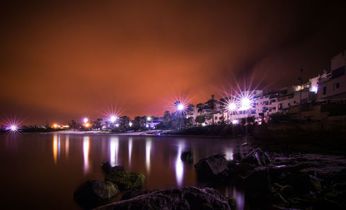 Illuminated city by river against sky at night