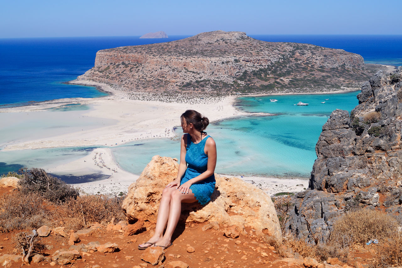 YOUNG WOMAN SITTING ON ROCK BY SEA AGAINST CLEAR SKY