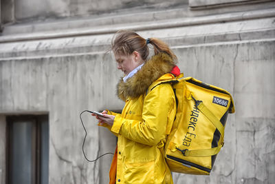 Side view of woman using mobile phone while standing against yellow wall