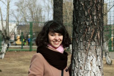 Portrait of smiling young woman standing by tree trunk at park