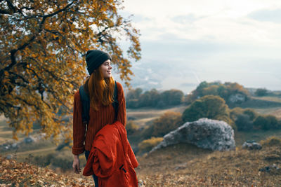 Young woman looking away while standing by tree against sky