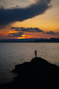 Silhouette man standing on rock by sea against sky during sunset