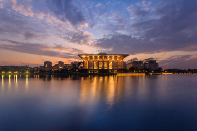 Illuminated buildings by lake against sky during sunset