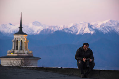 Man using camera while sitting against snowcapped mountains