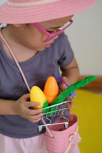 High angle view of cute girl holding small shopping cart with toys while standing against wall