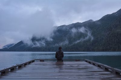 Rear view of man sitting on jetty over lake against mountain