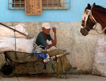 Man sitting on wheelbarrow with horse by wall