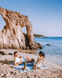 People sitting on rock by sea against clear sky