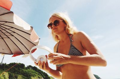Low angle view of woman holding headphones while wearing sunglasses against sky