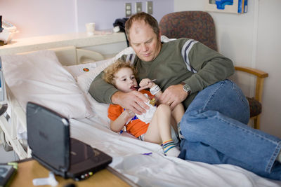 Dad cuddlse with toddler in hospital bed as toddler eats snack.