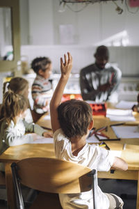 Rear view of boy raising hand while sitting on chair in classroom at kindergarten