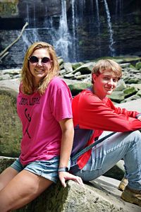 Portrait of couple smiling while sitting on rock against waterfall