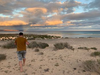 Rear view of man walking at beach against cloudy sky during sunset