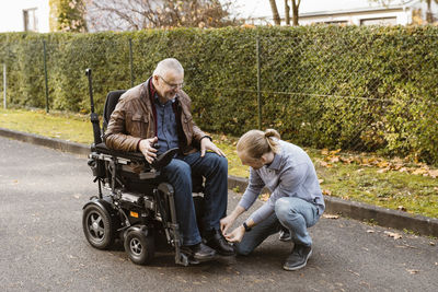 Young man kneeling while tying shoelace of father with disability sitting in motorized wheelchair