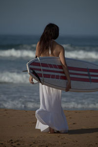 Rear view of woman standing at beach while holding surfboard against clear sky