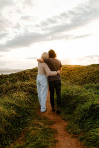 Back view blond woman and man embracing each other and looking away while standing on grassy cliff at sundown in aviles, spain