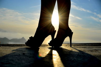 Low section of woman wearing high heels standing on road during sunset