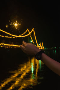 Cropped hand holding illuminated sparkler over river at night