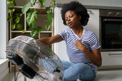 Exhausted african woman sitting in front of electric fan at home, using air cooler indoors