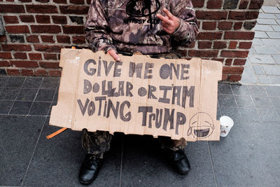 Low section of beggar with sign sitting by brick wall