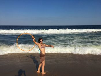 Side view of young woman with plastic hoop standing at beach against clear sky