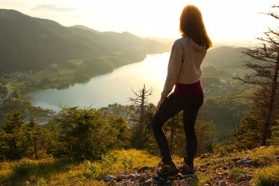 Rear view of young woman looking at lake while standing on mountain against sky during sunset