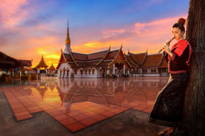 Young woman playing flute while standing against temple during sunset