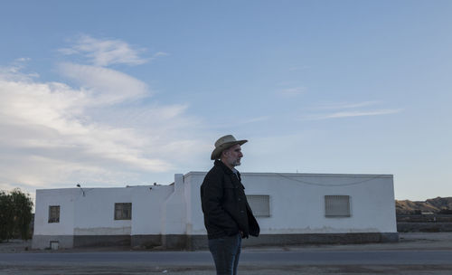 Adult man in cowboy hat standing in front of white building against sky