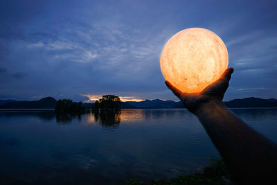 Cropped hand holding illuminated lighting equipment over lake against cloudy sky at dusk