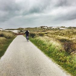 Rear view of man riding bicycle on road leading towards cloudy sky