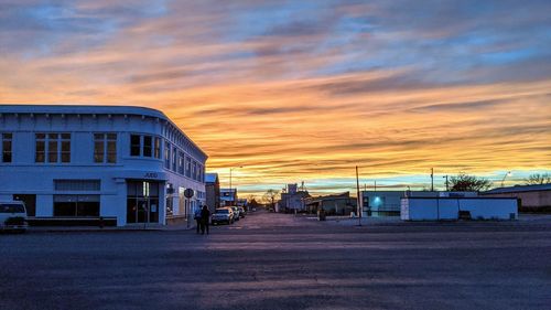 Road by buildings against sky during sunset