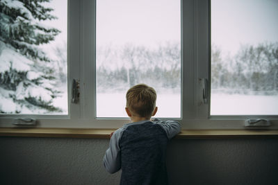 Rear view of boy looking through window while standing at home during winter