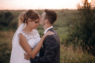 Side view of husband kissing on wife forehead against sky during sunset