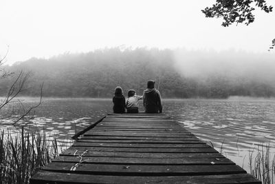 Rear view of mother with daughters sitting on pier at lake during foggy weather