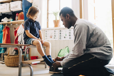 Side view of male teacher helping boy in wearing shoe at kindergarten