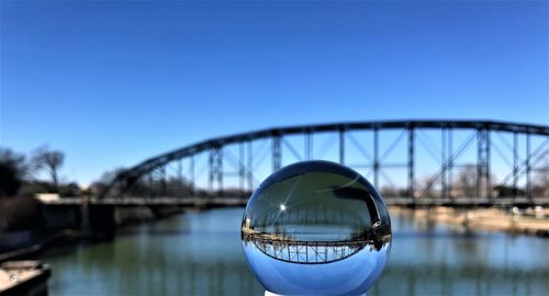 Reflection of arch bridge in crystal ball by river against clear blue sky