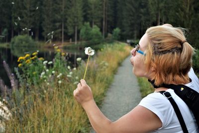 Side view of woman blowing dandelion against trees