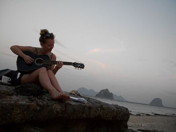 Full length of woman playing guitar while sitting on rock at beach