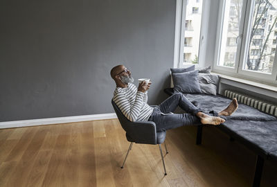 Matureman with earphones sitting at window, drinking coffee barefoot