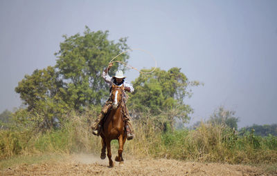 Cowboy holding rope riding horse against sky